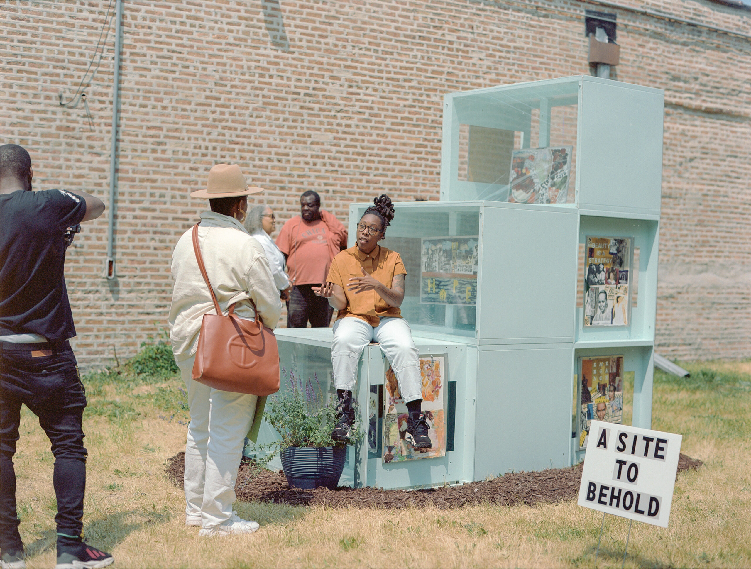 Artist is seated on top of art work that is placed outside in front of a brick wall. Artist is pictured in discussion with art viewers and a photographer.